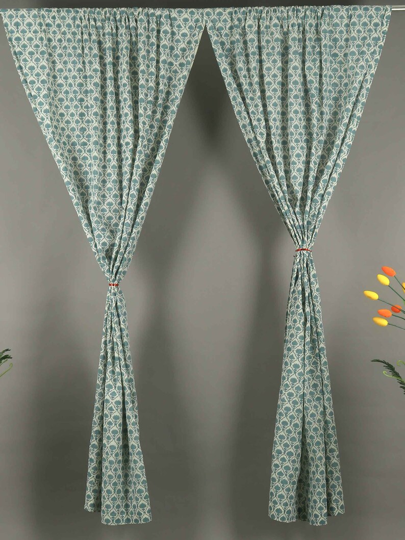 May include: A pair of floor-length curtains featuring a repeating teal and white floral pattern. The fabric is gathered at the center of each panel with a thin red tie, creating a draped effect that hangs from a rod at the top of the panels.