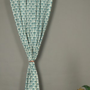 May include: A single curtain panel featuring a repeating teal and white floral block print pattern. The fabric is gathered at the center with a thin orange tie, creating a draped effect while hanging from a silver metal rod at the top.