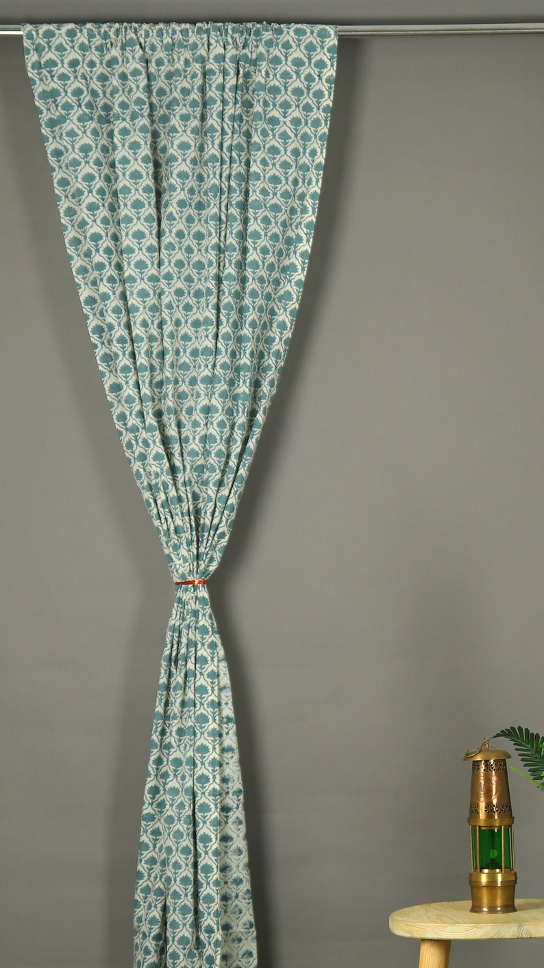 May include: A single curtain panel featuring a repeating teal and white floral block print pattern. The fabric is gathered at the center with a thin orange tie, creating a draped effect while hanging from a silver metal rod at the top.