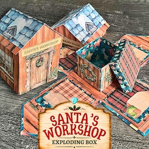 May include: A paper craft exploding box designed as Santa's workshop, featuring a house-shaped lid with a blue shingled roof, wood-textured walls, a wreath on the door, and interior panels decorated with red plaid patterns and star-filled blue borders.