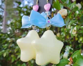 May include: A pair of star-shaped earrings with a light yellow glow, featuring a blue bow and pink heart charms. The earrings are hanging from a black display card.