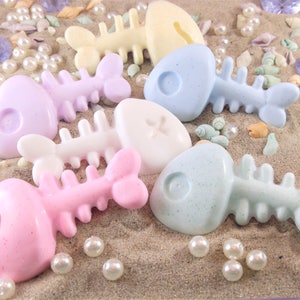 May include: Five colorful fish-shaped soaps in pastel shades of pink, blue, yellow, and purple. The soaps are arranged on a sandy surface with seashells and pearls.