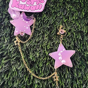 May include: A pair of star-shaped earrings with a gold chain and a crescent moon charm. The earrings are lavender and pink with pearl accents. The image also includes a pink and purple sticker with the text "Plushi Kawaii".