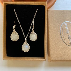 May include: A silver necklace and earrings set with a white moonstone pendant and matching earrings. The jewelry is displayed in a brown box with a black interior.