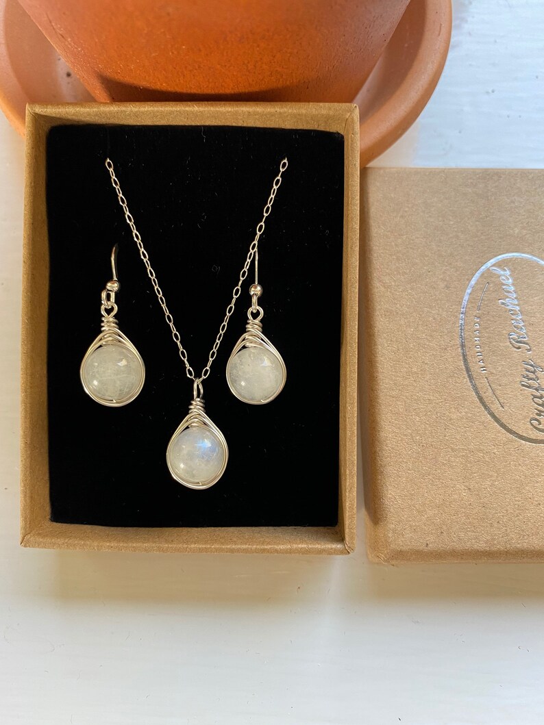 May include: A silver necklace and earrings set with a white moonstone pendant and matching earrings. The jewelry is displayed in a brown box with a black interior.