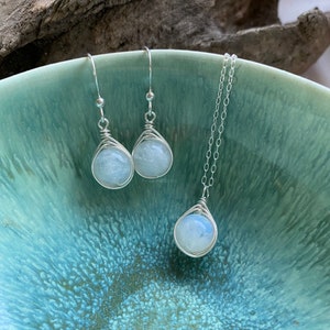 May include: A set of silver wire wrapped moonstone earrings and a matching necklace. The stones are a pale blue color and have a pearlescent sheen.