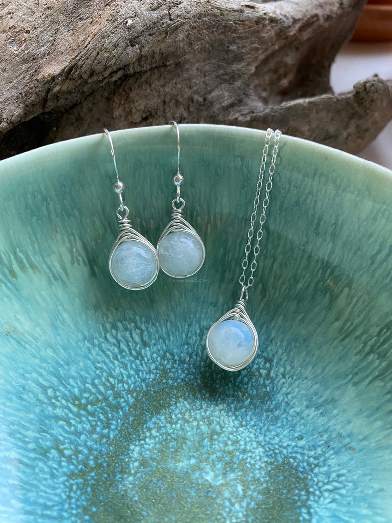 May include: A set of silver wire wrapped moonstone earrings and a matching necklace. The stones are a pale blue color and have a pearlescent sheen.