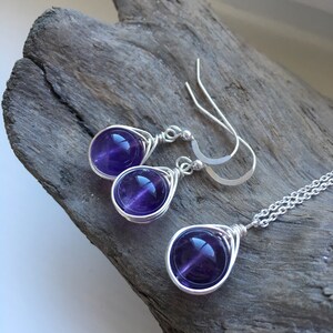 May include: A set of amethyst gemstone earrings and a necklace. The earrings and necklace feature a teardrop-shaped design with a silver wire wrapped around each gemstone.