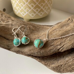May include: A set of turquoise gemstone jewelry featuring a necklace and earrings. The necklace has a silver chain and a turquoise pendant wrapped in silver wire. The earrings are silver with turquoise stones wrapped in silver wire.