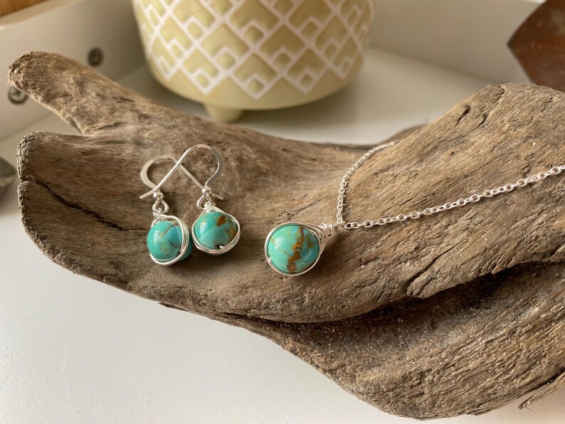 May include: A set of turquoise gemstone jewelry featuring a necklace and earrings. The necklace has a silver chain and a turquoise pendant wrapped in silver wire. The earrings are silver with turquoise stones wrapped in silver wire.