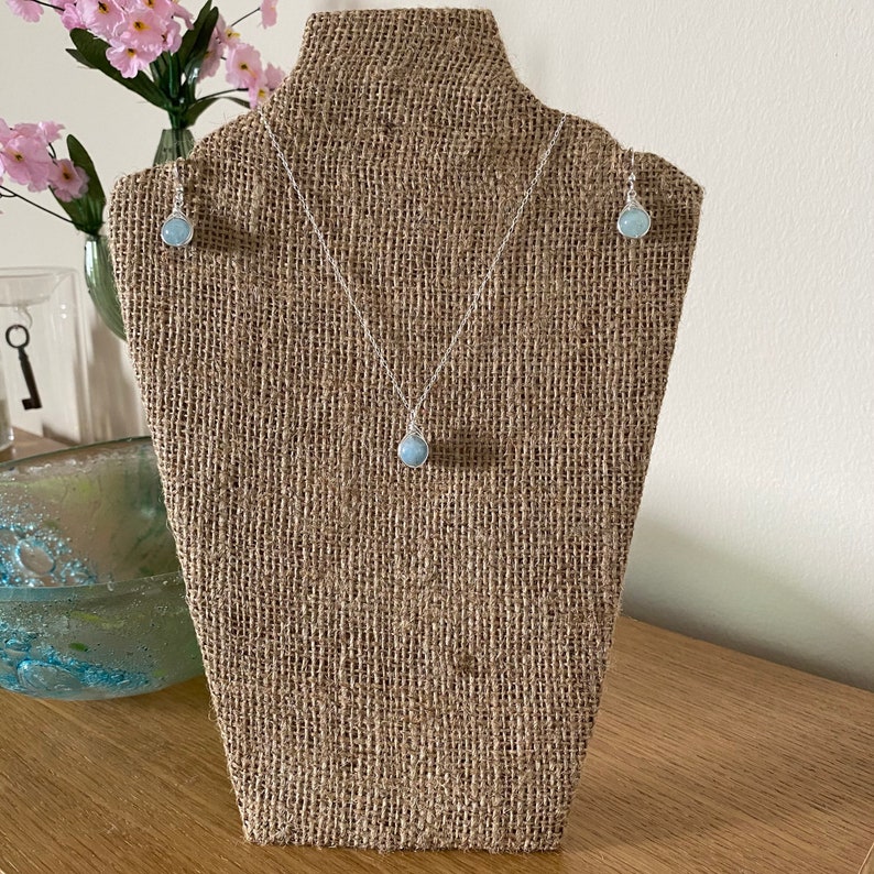 K&ouml;nnte beinhalten: Eine zarte silberne Halskette und passende Ohrringe mit einem hellblauen Edelstein-Anh&auml;nger. Der Schmuck ist auf einer Jute-Puppe ausgestellt.