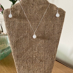 May include: A white gemstone necklace and earrings set on a burlap display stand. The necklace and earrings feature a round, white gemstone pendant and matching earrings.
