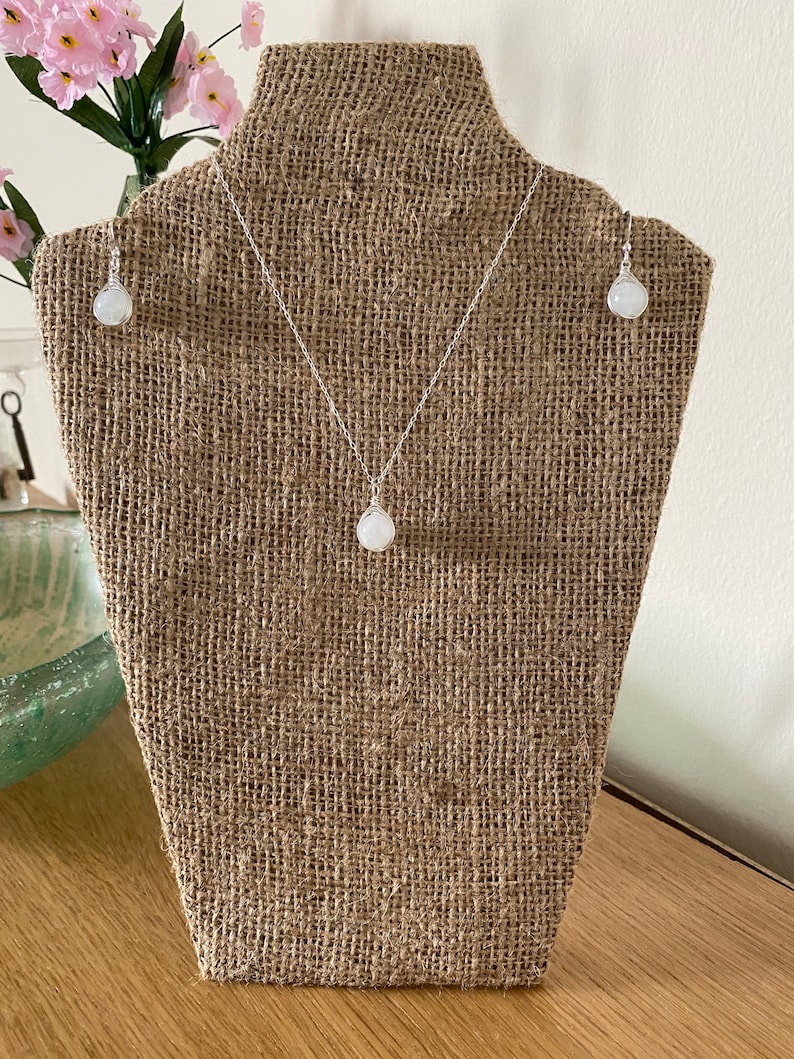 May include: A white gemstone necklace and earrings set on a burlap display stand. The necklace and earrings feature a round, white gemstone pendant and matching earrings.