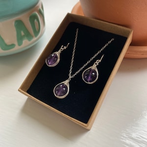 May include: A set of amethyst gemstone jewelry, including a necklace and earrings. The amethyst stones are wrapped in silver wire and are a deep purple color. The jewelry is displayed in a small, brown box with a black velvet lining.
