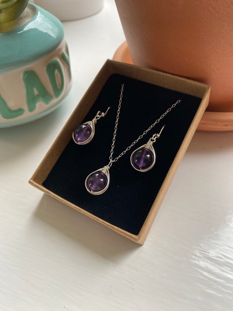 May include: A set of amethyst gemstone jewelry, including a necklace and earrings. The amethyst stones are wrapped in silver wire and are a deep purple color. The jewelry is displayed in a small, brown box with a black velvet lining.