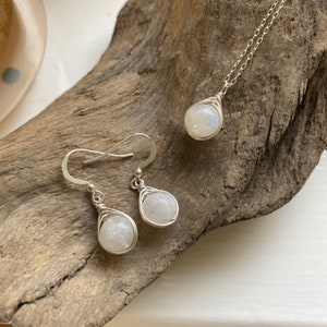 May include: A set of silver jewelry featuring moonstone earrings and a necklace. The stones are wrapped in silver wire and have a pearlescent white color.