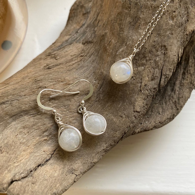 May include: A set of silver jewelry featuring moonstone earrings and a necklace. The stones are wrapped in silver wire and have a pearlescent white color.