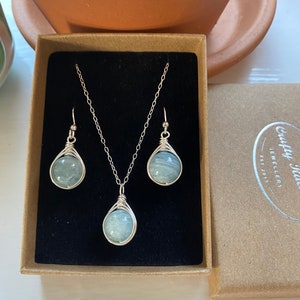 K&ouml;nnte beinhalten: Ein silbernes Halsketten- und Ohrring-Set mit einem hellblauen Edelstein-Anh&auml;nger und passenden Ohrringen. Der Schmuck ist in einer braunen Schachtel mit dem Text "Crafty Jules Jewellery Est 2013" ausgestellt.