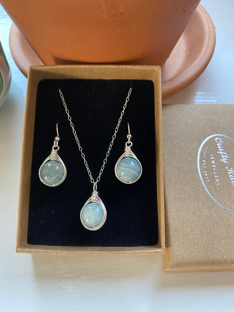 K&ouml;nnte beinhalten: Ein silbernes Halsketten- und Ohrring-Set mit einem hellblauen Edelstein-Anh&auml;nger und passenden Ohrringen. Der Schmuck ist in einer braunen Schachtel mit dem Text "Crafty Jules Jewellery Est 2013" ausgestellt.