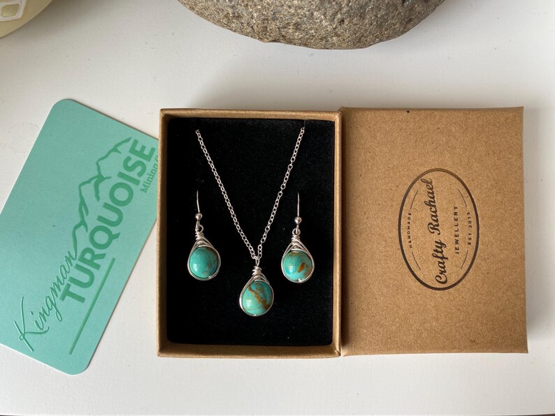 May include: A turquoise gemstone necklace and earrings set in a brown box. The necklace and earrings are made with silver wire and feature a turquoise stone. The box has the text "Crafty Rachael Jewellery" printed on it.