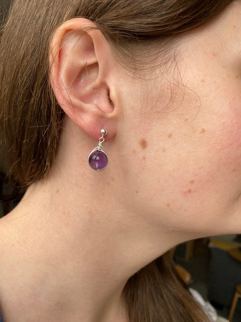 May include: A pair of silver earrings with a purple amethyst stone. The earrings are simple and elegant, perfect for everyday wear.