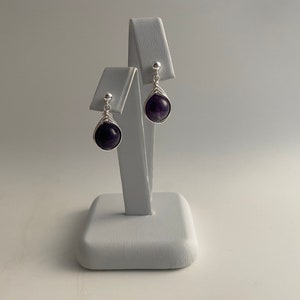 May include: A pair of silver dangle earrings with amethyst gemstones. The earrings are on a white display stand.
