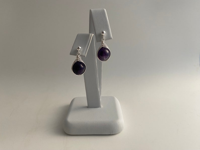 May include: A pair of silver dangle earrings with amethyst gemstones. The earrings are on a white display stand.