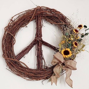 May include: A rustic peace sign wreath made of woven brown branches, adorned with sunflowers, dried grasses, and a burlap bow. The wreath is hanging on a white wall, creating a natural and welcoming aesthetic.