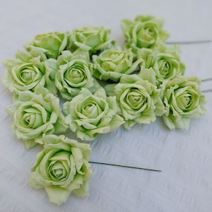 May include: A collection of light green paper roses with wire stems. The roses are in full bloom, with multiple layers of petals. The flowers are arranged on a white textured surface, suitable for crafts or floral arrangements.