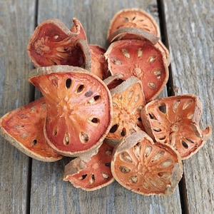 May include: A pile of dried bael fruit slices, showcasing a reddish-orange hue with a textured surface and a network of seed cavities. The fruit is arranged on a weathered, gray wooden surface, highlighting the natural product.