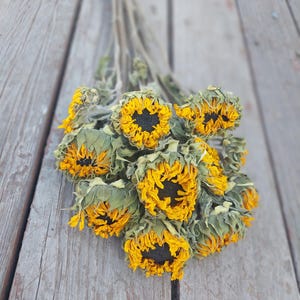 May include: A bouquet of dried sunflowers with yellow petals and dark centers. The flowers are clustered together, with some of the green leaves still attached. The background is a weathered wooden surface.