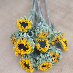 May include: A bundle of dried sunflowers with yellow petals and dark centers. The flowers have green and gray stems and leaves. The arrangement is set against a light brown wooden surface.