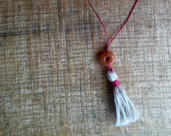 Puede incluir: Un collar de cordón rojo con un colgante de piedra marrón y un borla blanca.