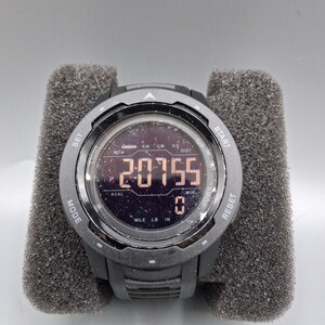 May include: A black digital sports watch with a round face, displaying the number 20755. The watch has multiple buttons and is set in a gray foam holder. The watch face shows various metrics.