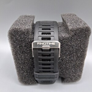 May include: Black wristwatch with a textured black band and a silver clasp. The watch band has a rectangular design with the word "FINOTIME" printed on the clasp. The watch is resting on a gray foam surface.