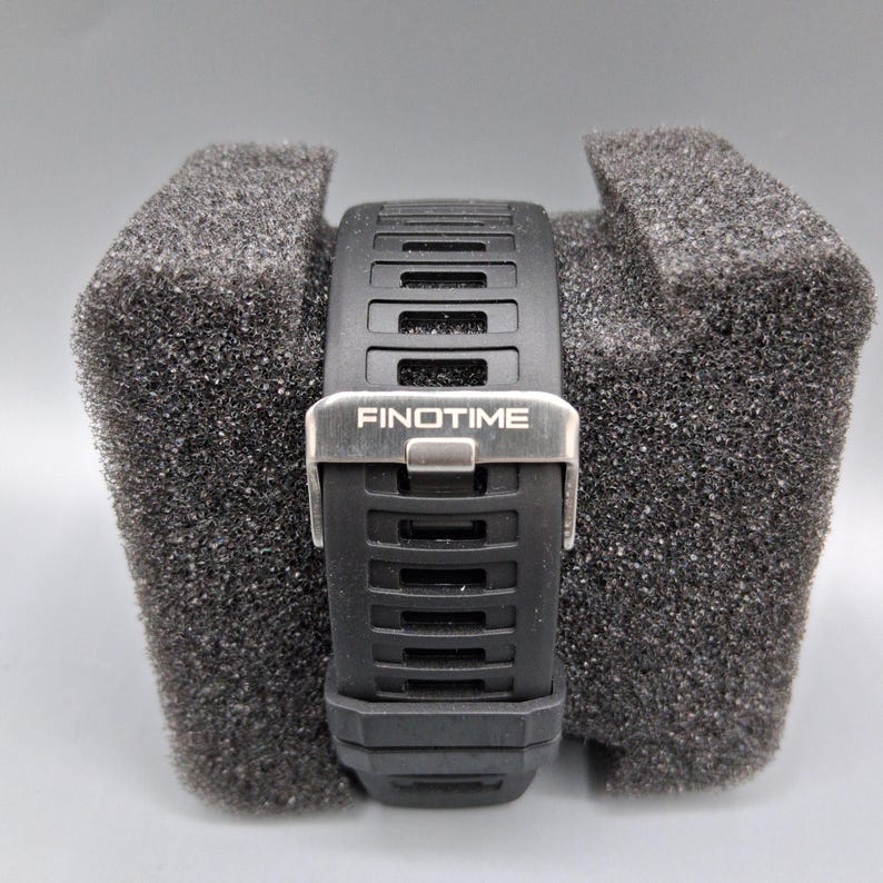 May include: Black wristwatch with a textured black band and a silver clasp. The watch band has a rectangular design with the word "FINOTIME" printed on the clasp. The watch is resting on a gray foam surface.