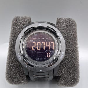 May include: A black digital sports watch with a round face, displaying the number "20747". The watch has multiple buttons and displays units like "MILE", "LB", and "IN". It is set on a gray foam surface.