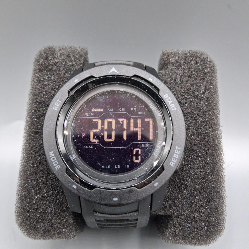 May include: A black digital sports watch with a round face, displaying the number "20747". The watch has multiple buttons and displays units like "MILE", "LB", and "IN". It is set on a gray foam surface.
