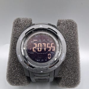 May include: A black digital sports watch with a round face, displaying the number 20755. The watch has multiple buttons and is set in a gray foam holder. The watch face shows various metrics.
