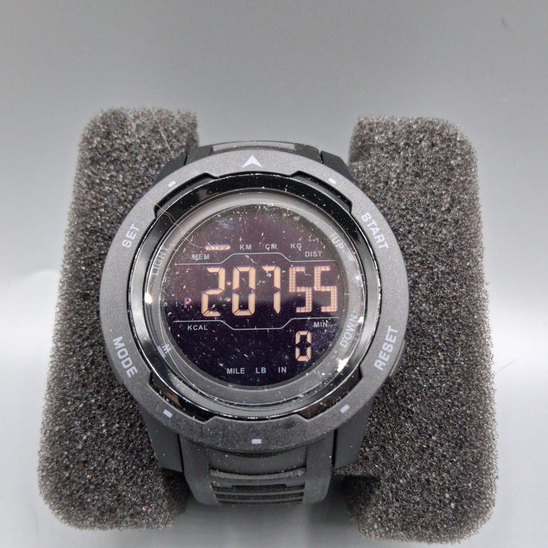 May include: A black digital sports watch with a round face, displaying the number 20755. The watch has multiple buttons and is set in a gray foam holder. The watch face shows various metrics.