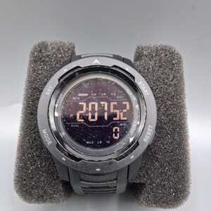 May include: A black digital sports watch with a round face, displaying numbers and symbols. The watch has multiple buttons and is held in a gray foam holder. The watch face shows the numbers "20752".