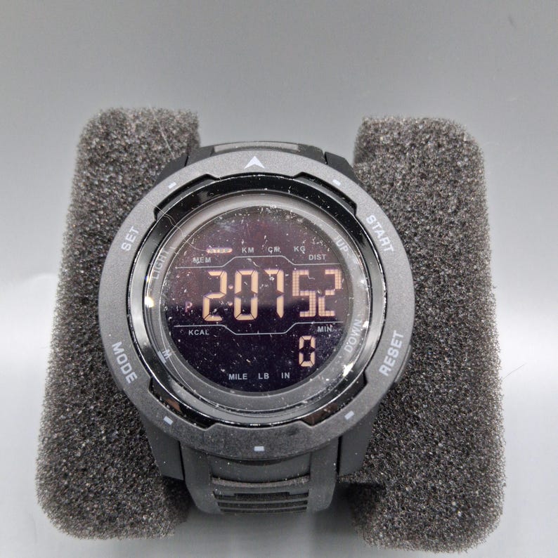 May include: A black digital sports watch with a round face, displaying numbers and symbols. The watch has multiple buttons and is held in a gray foam holder. The watch face shows the numbers "20752".