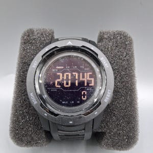May include: A black digital sports watch with a round face, displaying the number "20745". The watch has multiple buttons and displays units like "KCAL", "MILE", "LB", and "IN". The watch is set on a gray foam.