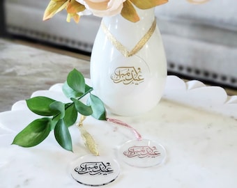 Può includere: Due ornamenti rotondi in acrilico trasparente con calligrafia araba dorata e rosa che recita "Eid Mubarak". Gli ornamenti sono appesi a nastri dorati e rosa. Gli ornamenti sono su una superficie bianca con un vaso di peonie rosa e pesca sullo sfondo.