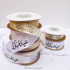 Peut inclure: Rouleaux de ruban blanc et beige avec une calligraphie arabe dorée qui se lit "Eid Mubarak" en anglais. Le ruban est sur une surface en marbre blanc.