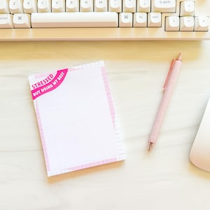 May include: A pink and white notepad with the text "STRESSED BUT DOING MY BEST" sits on a white surface. A pink pen and a white computer mouse are next to the notepad. A white keyboard is in the background.