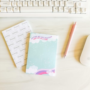 May include: A light blue notepad with the text "GETTING MY LIFE TOGETHER ONE LIST AT A TIME" and a rainbow design. A Post-it note pad and a pink pen are also on the white surface, along with a white keyboard and a computer mouse.