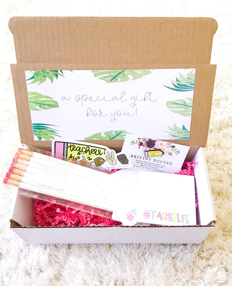 May include: A white cardboard box with pink crinkle paper inside. The box contains a set of pencils, a sticker that says "Teacher" and a sticker that says "#TeacherLife". The box also contains a business card for Brittny Nguyen.