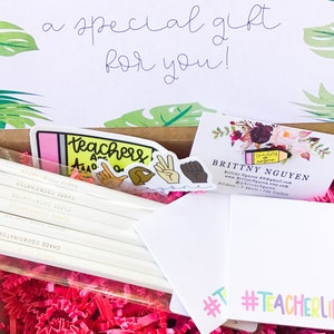 May include: A gift box filled with pink crinkle paper and teacher-themed gifts. The box includes a set of pencils with the text "Chaos Coordinator" printed on them, a sticker that says "Teachers are awesome", a sticker with a fist raised in the air, and a business card for Brittny Nguyen. The business card includes the text "Teachers are awesome" and the website address www.BrittayNguyen.etsy.com. The box also includes a white card with the hashtag #TeacherLife.