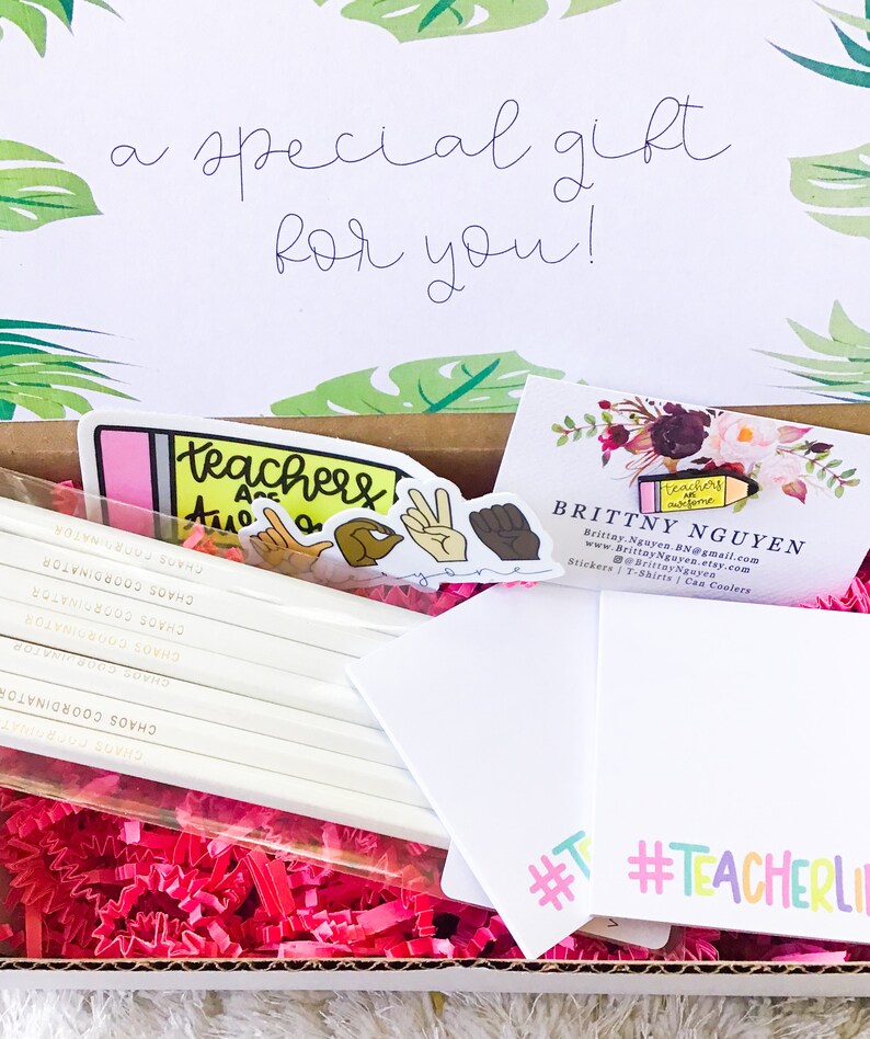 May include: A gift box filled with pink crinkle paper and teacher-themed gifts. The box includes a set of pencils with the text "Chaos Coordinator" printed on them, a sticker that says "Teachers are awesome", a sticker with a fist raised in the air, and a business card for Brittny Nguyen. The business card includes the text "Teachers are awesome" and the website address www.BrittayNguyen.etsy.com. The box also includes a white card with the hashtag #TeacherLife.